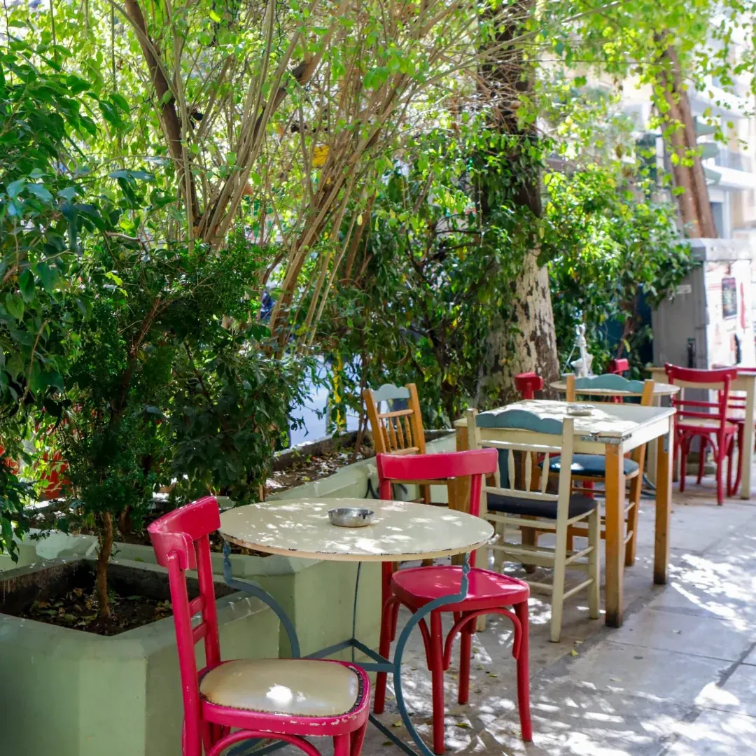 Κόκκινες καρέκλες μπιστρό και μεταλλικά τραπεζάκια στη σκιά των δέντρων, στο πεζοδρόμιο της Εμμ. Μπενάκη.