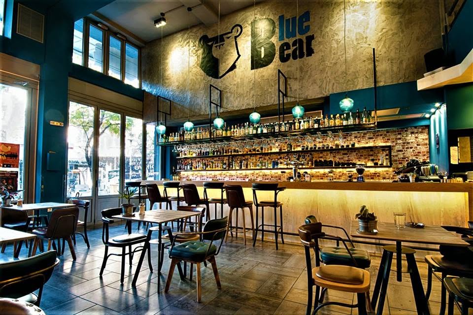 The Bluehaus bar in Exarcheia — exposed-brick wall with the hand-painted Blue Bear logo, backlit bottle shelves and teal glass pendant lights