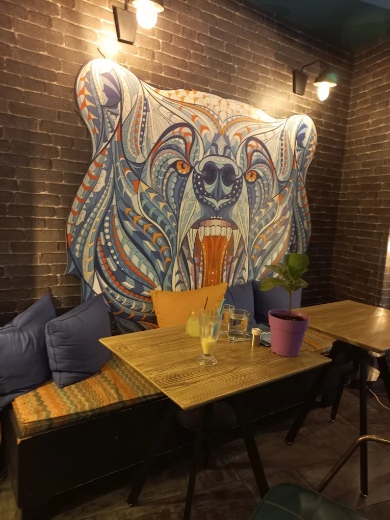 A corner booth beneath the hand-painted Blue Bear mural — an intricately patterned bear in blues, teals and orange on exposed brick, with cushioned bench seating and a fresh juice on the table
