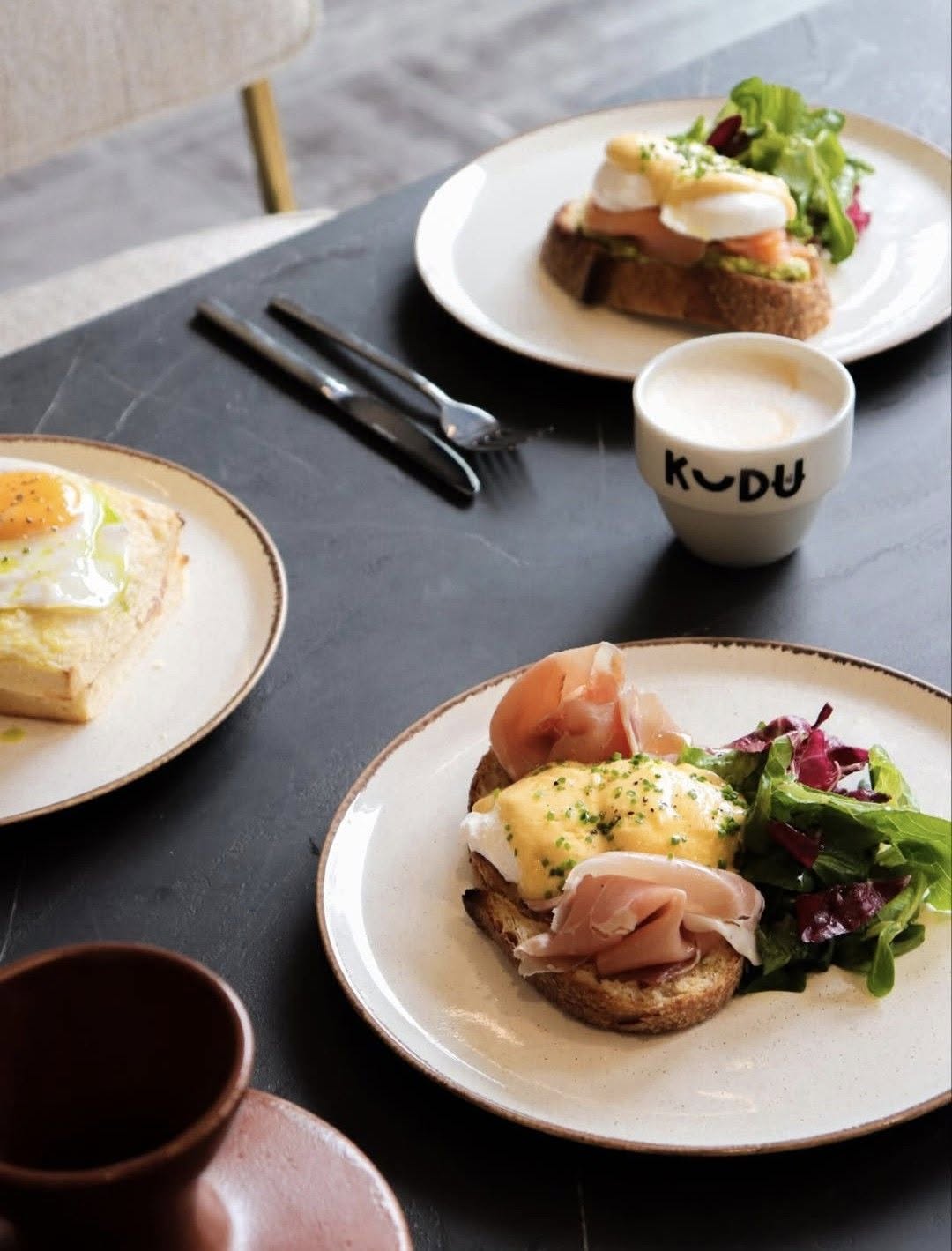 Bluehaus brunch — eggs benedict with prosciutto and dressed greens, a second poached-egg toast, and a KUDU cup of cappuccino on a dark stone table