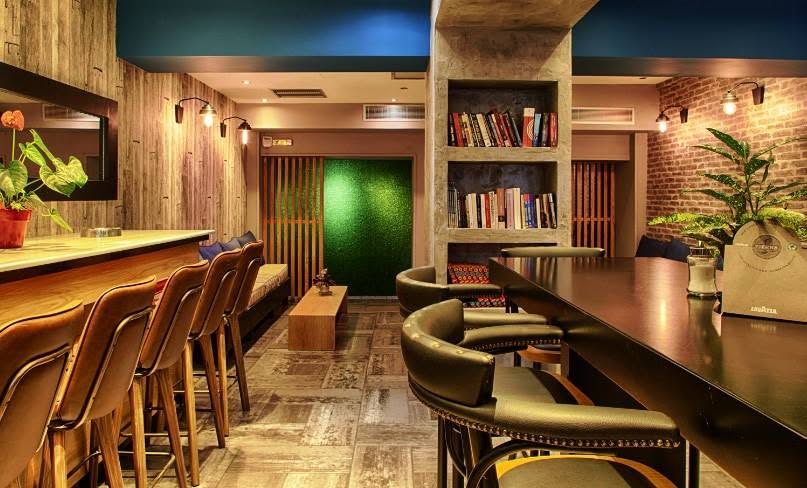 The Bluehaus lounge and library — wood-panelled walls, an exposed-brick nook, bookshelves, a moss panel and leather bar stools under a petrol-blue ceiling