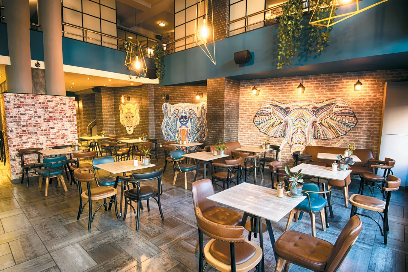 The main room — exposed brick walls with patterned lion, bear and elephant murals, leather and petrol-blue chairs, reclaimed-wood tables and geometric brass pendant lights